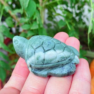 Peut inclure: Petite figurine de tortue sculptée, vert et blanc. La tortue est tenue dans une main, avec un arrière-plan vert flou. La figurine présente un motif de carapace détaillé et une finition lisse et polie.