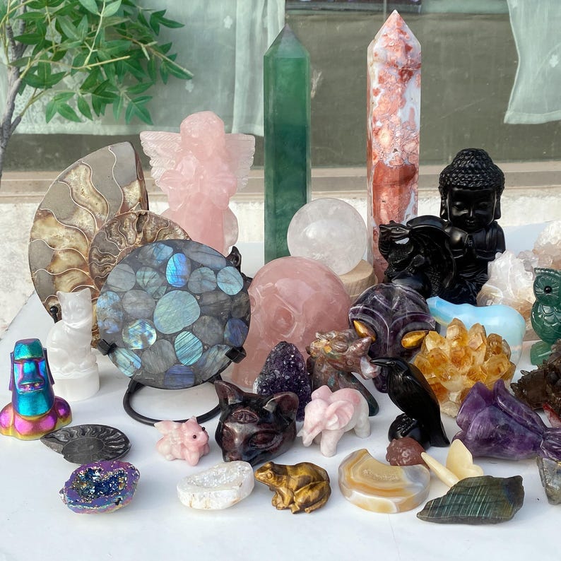 Pode incluir: Uma cole&ccedil;&atilde;o de v&aacute;rios cristais e pedras preciosas, incluindo um anjo de quartzo rosa, uma esfera de labradorita e uma est&aacute;tua de Buda negra. Outros itens incluem um obelisco verde, uma caveira rosa e figuras de animais.