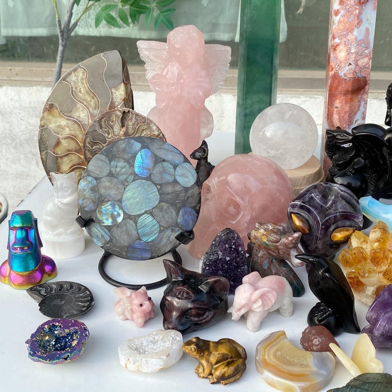 Pode incluir: Uma cole&ccedil;&atilde;o de v&aacute;rias esculturas de pedras preciosas e cristais. Inclui um anjo de quartzo rosa, uma esfera de labradorita, uma bola de cristal de quartzo e figuras de animais como um gato, um elefante e um sapo. A paleta de cores inclui tons de rosa, roxo e iridescente.
