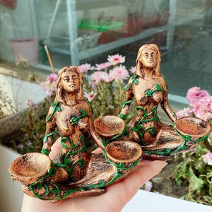 May include: Two bronze-colored mermaid figurines with green vine accents. Each mermaid holds two small, round bowls. The figurines are detailed with flowing hair and seated poses, ideal for decorative purposes.