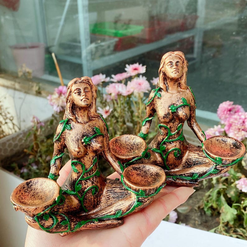 May include: Two bronze-colored mermaid figurines with green vine accents. Each mermaid holds two small, round bowls. The figurines are detailed with flowing hair and seated poses, ideal for decorative purposes.