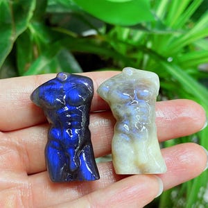 Könnte beinhalten: Zwei geschnitzte Torso-Skulpturen aus Edelsteinen. Eine ist tiefblau mit schillernden Reflexen, die andere hat eine blasse, cremefarbene Farbe mit subtilen Blautönen. Die Skulpturen werden in einer Hand vor einem Hintergrund aus grünem Laub gehalten.