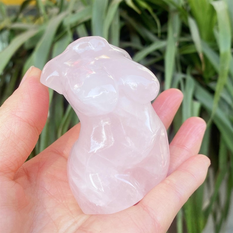 May include: A pink rose quartz torso sculpture. The carved figurine depicts a stylized female form. The stone has a translucent quality, with subtle variations in colour. The sculpture is held in a hand, with green foliage in the background.