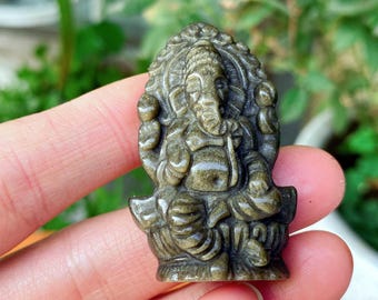 Puede incluir: Una detallada talla de piedra verde oliva del dios hindú Ganesha. La escultura representa a Ganesha sentado, con intrincados detalles en la cabeza de elefante, múltiples brazos y un tocado ornamentado. La talla se sostiene en una mano.