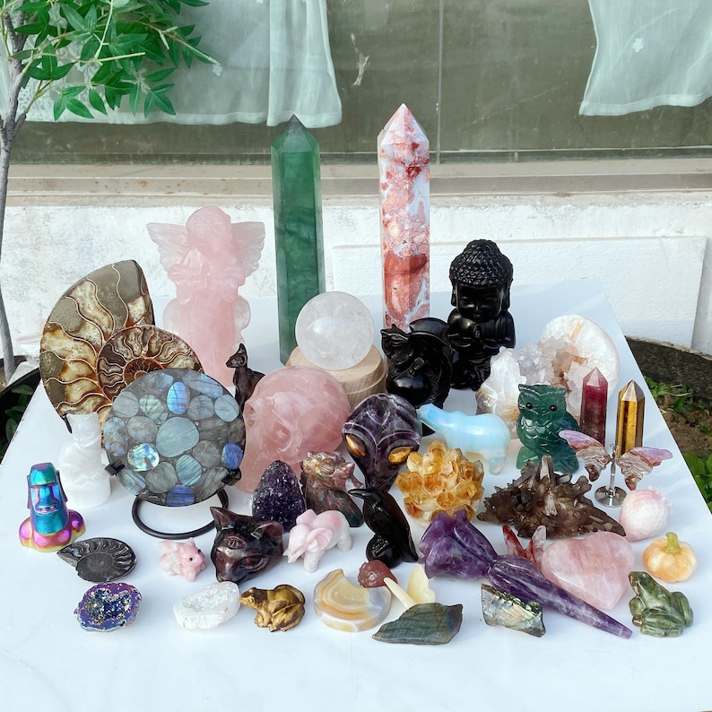Pode incluir: Uma cole&ccedil;&atilde;o de v&aacute;rias esculturas de pedras preciosas e cristais. Os itens incluem um anjo rosa, uma est&aacute;tua de Buda preta, uma esfera de quartzo transparente e v&aacute;rias torres de cristal. Outras esculturas incluem animais, caveiras e uma geodo.