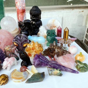 Pode incluir: Uma cole&ccedil;&atilde;o de estatuetas de pedras preciosas esculpidas e cristais. Inclui uma est&aacute;tua de Buda preta, uma coruja verde, um corvo preto e v&aacute;rias forma&ccedil;&otilde;es de cristal em tons de rosa, roxo e dourado. H&aacute; uma variedade de formas e tamanhos.