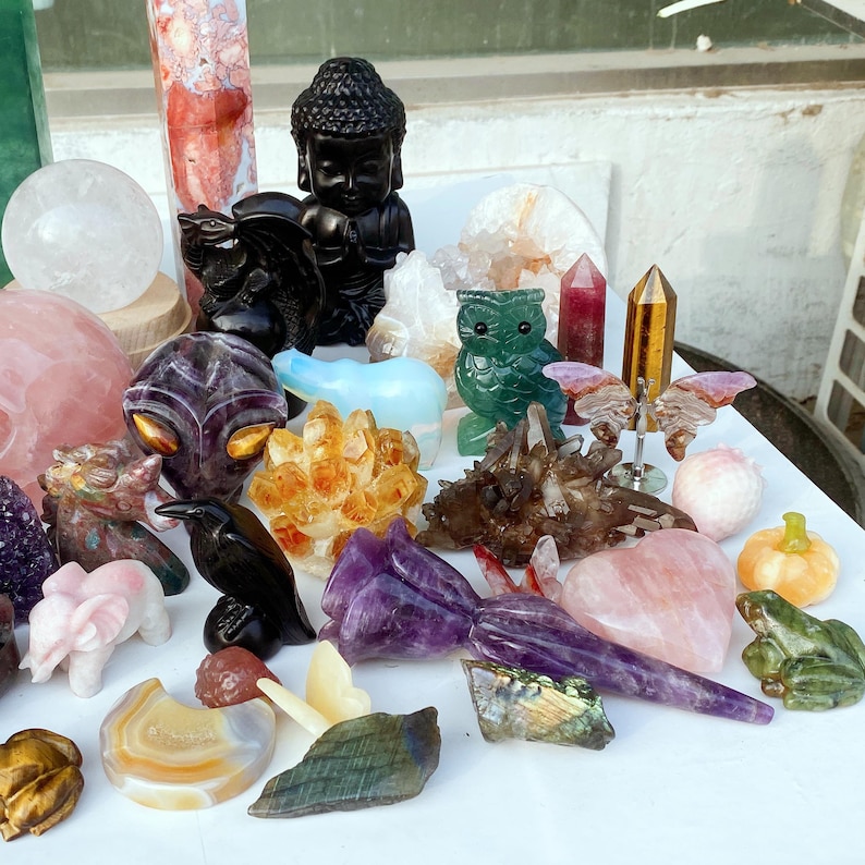 Pode incluir: Uma cole&ccedil;&atilde;o de estatuetas de pedras preciosas esculpidas e cristais. Inclui uma est&aacute;tua de Buda preta, uma coruja verde, um corvo preto e v&aacute;rias forma&ccedil;&otilde;es de cristal em tons de rosa, roxo e dourado. H&aacute; uma variedade de formas e tamanhos.