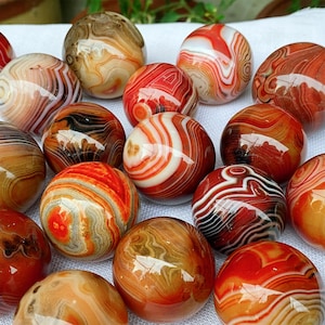 Puede incluir: Una colección de piedras de ágata pulidas en varios tonos de rojo, naranja, marrón y blanco. Las piedras tienen una superficie lisa y brillante y muestran intrincados patrones de bandas. Las piedras esféricas están dispuestas juntas.