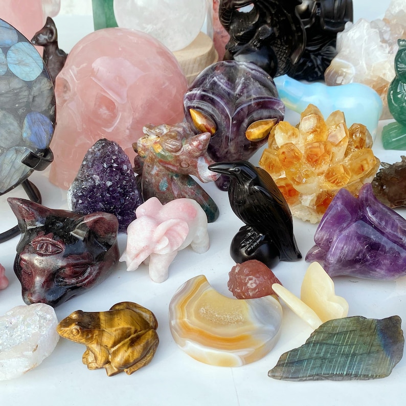 Pode incluir: Uma cole&ccedil;&atilde;o de estatuetas esculpidas em pedras preciosas, incluindo um cr&acirc;nio rosa, um corvo preto, um elefante rosa e um sapo amarelo. Outras pedras incluem ametista, citrino e &aacute;gata, mostrando uma variedade de cores e texturas.