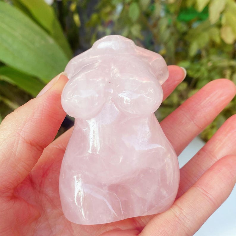 May include: A pink rose quartz figurine shaped like a torso. The polished sculpture features rounded forms representing the breasts and abdomen. The translucent stone has subtle colour variations. The figurine is held in a hand.