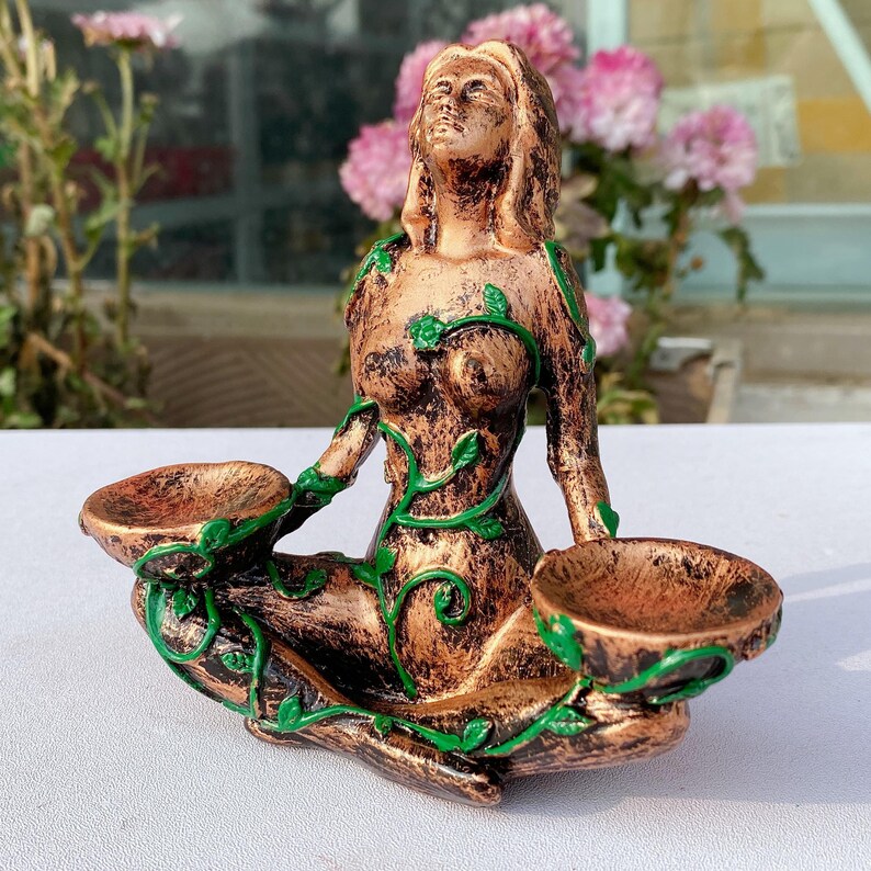 May include: A bronze-colored figurine of a seated woman with outstretched arms, holding two shallow bowls. Green vines with leaves adorn the figure's body. The statue has a weathered appearance and is set against a blurred background of pink flowers.