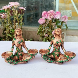 May include: Two bronze-colored, seated female figurines with green vine accents. Each holds two small, round bowls. The figurines are displayed on a white surface, with pink flowers in the background.