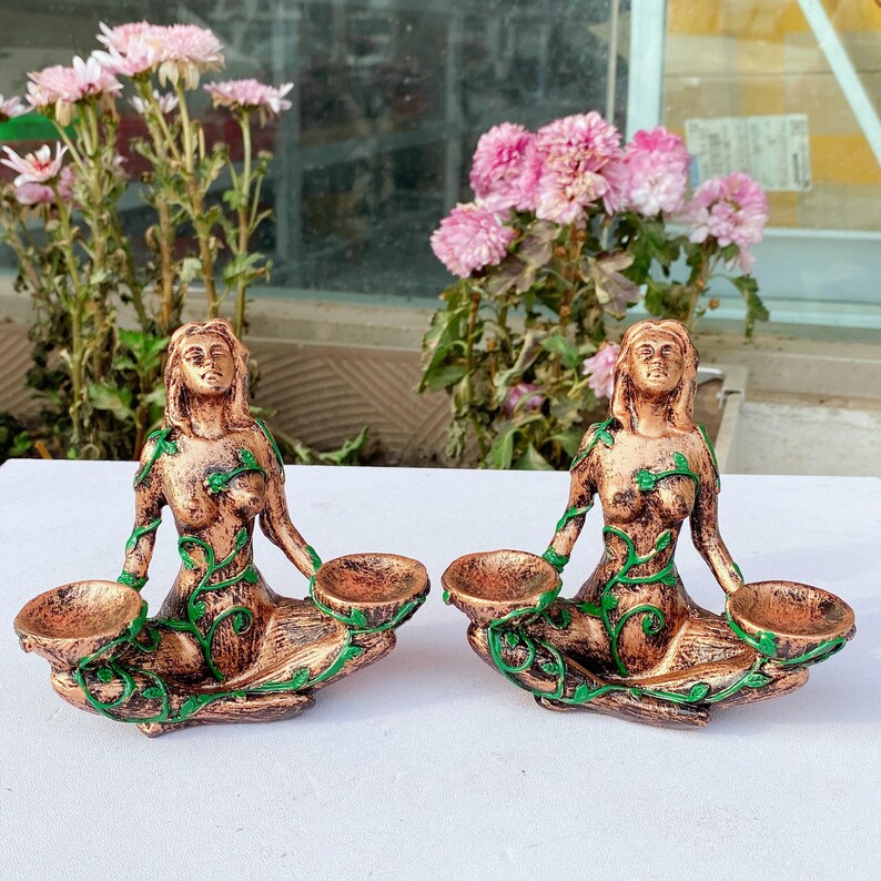 May include: Two bronze-colored, seated female figurines with green vine accents. Each holds two small, round bowls. The figurines are displayed on a white surface, with pink flowers in the background.