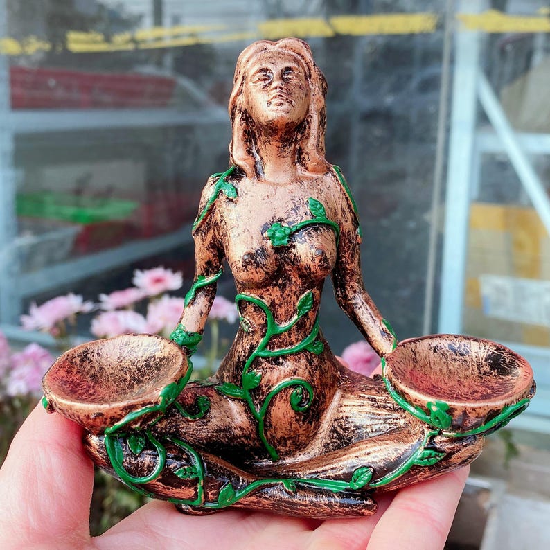 May include: A bronze-colored statue of a seated woman with long hair, holding two small bowls. Green vines with leaves are painted on the statue. The statue is held in a person's hand.