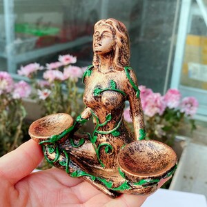 May include: A bronze-colored figurine of a seated woman with closed eyes, adorned with green vine-like accents. The figure holds two small bowls. The statue is approximately 6 inches tall and is likely a decorative item.