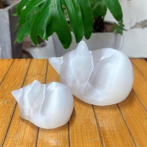 Puede incluir: Dos figuras de gato blancas en posición de dormir. El gato más grande está acurrucado, mientras que el más pequeño está en una pose similar. Las figuras están sobre una superficie de madera, con una planta verde al fondo. Los gatos son de un material liso y brillante.