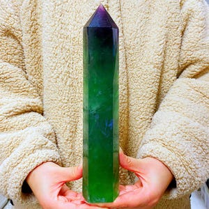 Può includere: Una punta di cristallo di fluorite lucidata e di forma allungata. Il cristallo passa da una punta viola scuro a sfumature di verde. I lati sfaccettati del cristallo sono visibili e la forma generale è quella di un obelisco lungo e snello.