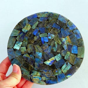 May include: Round coaster made of mosaic pieces of labradorite stone. The coaster features a mix of blue, green, and gray tones, with a polished surface. The mosaic design is arranged in a circular pattern, creating a decorative and functional item.
