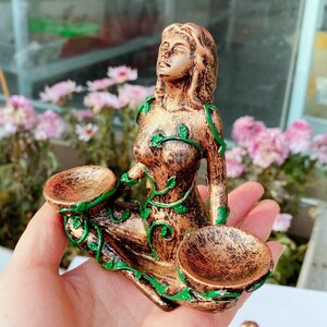 May include: A bronze-colored statue of a woman with closed eyes, adorned with green vine-like accents. The statue holds two small bowls. The woman's hair is shoulder-length, and she is wearing a dress. The statue is likely a decorative item.