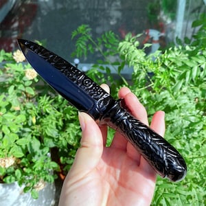 Puede incluir: Una daga negra con intrincadas tallas en la hoja y el mango. El mango presenta un diseño tejido. La daga se sostiene en una mano sobre un fondo de follaje verde.