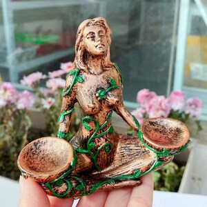 May include: A bronze-colored statue of a seated woman with long hair and closed eyes. Green vines with leaves are painted on the statue. The woman is holding two shallow bowls. The statue is likely a decorative item.