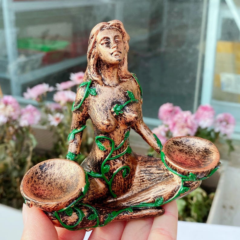 May include: A bronze-colored statue of a seated woman with long hair and closed eyes. Green vines with leaves are painted on the statue. The woman is holding two shallow bowls. The statue is likely a decorative item.