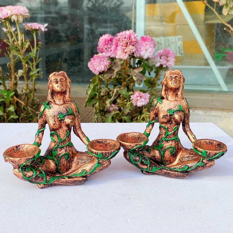 May include: Two bronze-colored statues of seated figures, each holding two small bowls. Green vine-like details adorn the figures. The statues are on a white surface, with pink flowers in the background.