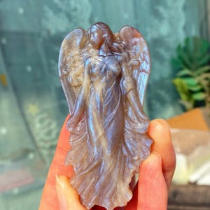 Könnte beinhalten: Eine geschnitzte Engelsfigur mit schillernden blauen und violetten Farbtönen. Der Engel hat detaillierte Flügel und fließende Gewänder. Die Skulptur wird in einer Hand gehalten, die die komplizierten Details der Steinschnitzerei zeigt.