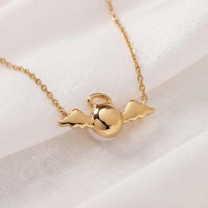 May include: A gold-colored necklace featuring a pendant with a round, polished center and small, stylized wings on either side. The necklace chain is delicate and also gold-toned, set against a white fabric background.