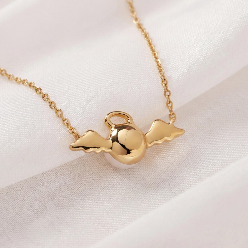 May include: A gold-colored necklace featuring a pendant with a round, polished center and small, stylized wings on either side. The necklace chain is delicate and also gold-toned, set against a white fabric background.