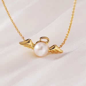 May include: A gold-colored necklace featuring a pearl pendant with angel wings. The pearl is round and white, and the wings are detailed with feather-like textures. The chain is delicate and also gold-colored, creating a simple yet elegant design.