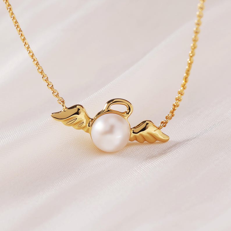 May include: A gold-colored necklace featuring a pearl pendant with angel wings. The pearl is round and white, and the wings are detailed with feather-like textures. The chain is delicate and also gold-colored, creating a simple yet elegant design.