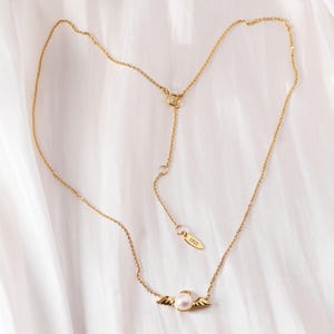 May include: A delicate gold-colored necklace with a pearl pendant and small wing accents. The necklace has an adjustable chain and a small tag with the number "3925". The jewelry is displayed on a white, textured surface.