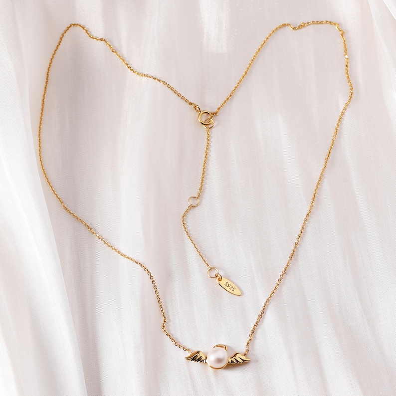 May include: A delicate gold-colored necklace with a pearl pendant and small wing accents. The necklace has an adjustable chain and a small tag with the number "3925". The jewelry is displayed on a white, textured surface.
