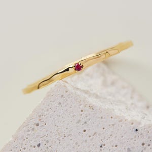May include: A gold ring with a textured band and a small, round, red gemstone. The ring is set against a neutral background, highlighting its simple elegance. The gemstone is a vibrant focal point.