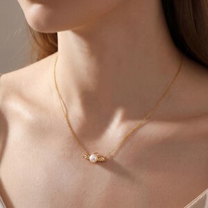 May include: A delicate gold necklace featuring a small, round pearl pendant. The pendant is framed by decorative gold accents. The necklace is shown on a person's neck, highlighting its elegant design and subtle sparkle.