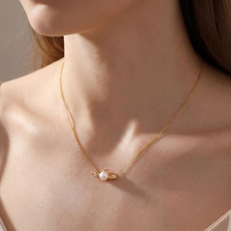 May include: A delicate gold necklace featuring a small, round pearl pendant. The pendant is framed by decorative gold accents. The necklace is shown on a person's neck, highlighting its elegant design and subtle sparkle.