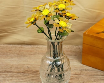 May include: A clear glass vase with a textured design holds a bouquet of yellow and green glass flowers. The flowers have yellow centers and translucent petals. The vase sits on a wooden surface, with a wooden box in the background.