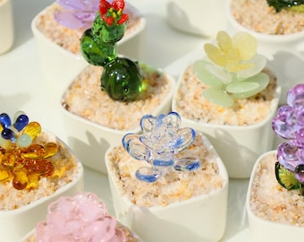 May include: A collection of colorful glass succulent sculptures in small white square pots filled with sand. The glass plants are various colors, including green, yellow, blue, pink, and purple. The sculptures are detailed and artistic.