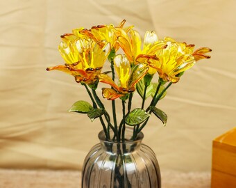 May include: A bouquet of yellow and orange glass flowers with green leaves in a gray glass vase. The flowers have a detailed, layered petal design. The vase has vertical ridges.