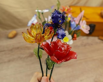 Puede incluir: Una mano sostiene un ramo de flores de cristal. El ramo incluye una rosa roja, un loto amarillo y un iris azul. En el fondo se ven otras flores de cristal, incluyendo mariposas y otras flores.