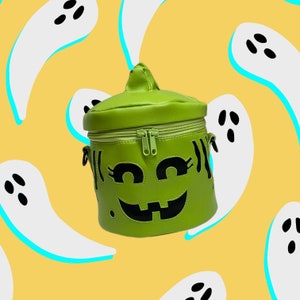 May include: A green pumpkin-shaped bag with a zipper closure and black eyes, nose, and mouth. The bag is on a yellow background with white ghost shapes.