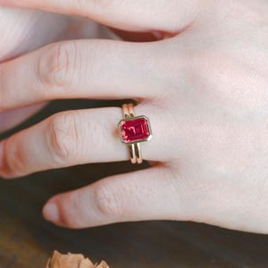 May include: A gold ring featuring a rectangular, emerald-cut red gemstone set in a smooth bezel setting. The band is thin and polished, showcasing a clean, metallic finish that complements the vibrant, deep red hue of the central stone.