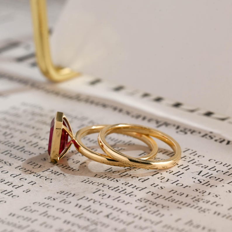 May include: A gold ring featuring a rectangular, deep red gemstone set in a minimalist metal frame. The band is thin and polished, showcasing a smooth, reflective surface that catches the light. The design is simple, focusing on the contrast between the vibrant red stone and the warm, metallic gold band.