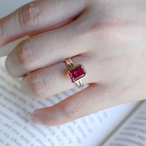 May include: A gold ring featuring a rectangular, emerald-cut, vibrant pink gemstone set in a smooth bezel setting. The band is thin and polished, showcasing a minimalist design that highlights the deep pink colour and faceted surface of the stone.