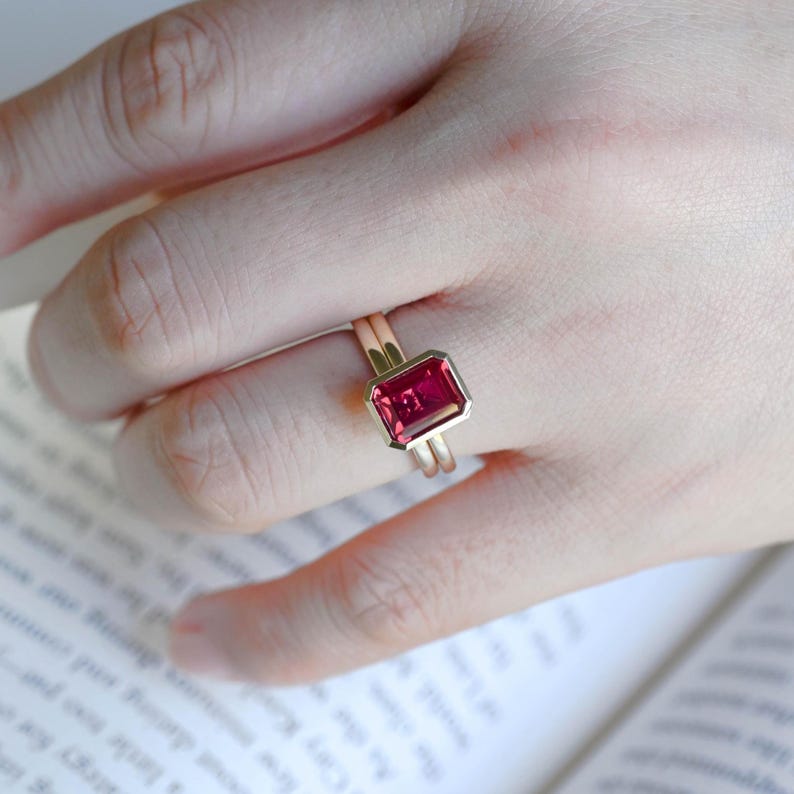 May include: A gold ring featuring a rectangular, emerald-cut, vibrant pink gemstone set in a smooth bezel setting. The band is thin and polished, showcasing a minimalist design that highlights the deep pink colour and faceted surface of the stone.