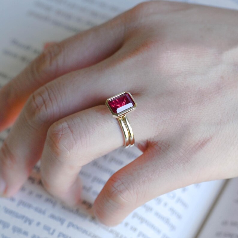 May include: A gold ring featuring a rectangular, emerald-cut deep red gemstone set in a smooth bezel setting. The band is thin and polished, showcasing a minimalist design that highlights the vibrant colour and clarity of the central stone.