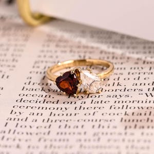 May include: A gold ring with a heart-shaped, dark red gemstone and a pear-shaped, clear gemstone. The ring is set on a vintage book page with printed text.