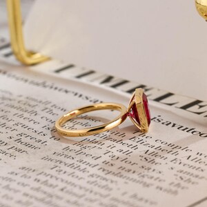 May include: A gold ring featuring a rectangular, faceted red gemstone set in a four-prong setting. The band is thin and polished, showcasing a vibrant crimson stone that reflects light. The ring is positioned on a surface with printed text.