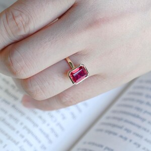 May include: A gold ring featuring a rectangular, emerald-cut vibrant pink gemstone set in a smooth bezel setting. The ring band is thin and polished, showcasing a clean, minimalist design that highlights the deep colour of the central stone.